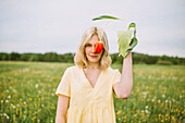 Delicate female covering eye with red tulip flower while standing in field and looking at camera