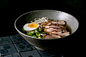 Asian style soup with rice noodles, grilled duck breast and boiled egg in ceramic bowl on dark background