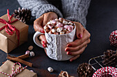 Holding mug of hot drink with marshmallows among Christmas gifts cones and cinnamon sticks