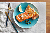 Baked salmon steak with lemon slice served on blue plate with fork and knife on white table