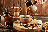 Adding freshly brewed coffee from cezve in metal cup placed on wooden table with vintage grinder and beans