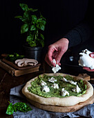Crop anonymous chef adding tasty Mozzarella cheese on pizza with pesto sauce and preparing lunch at dark table