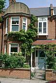 The exterior of a traditional pre war semi detached house covered in plants with a front garden and black metal gate