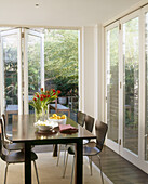A conservatory dining room with glass walls surrounding a modern dining table and chairs laden with food and flowers