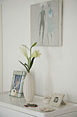 Weiße Vase mit Blumen und mehrere gerahmte Bilder auf Kommode unter einem Gemälde