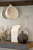 Large cups hanging from hooks above glassware items on wooden shelf