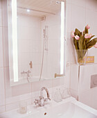 A detail of a modern bathroom white tiled walls wall mounted wash basin tap mirror reflecting shower over bath flower arrangement