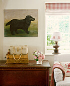 A detail of a country sitting room, wooden side cabinet, check roman blind at window, upholstered sofa, wicker hampers, painting of dog, flower arrangement, lamp