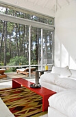 Modernes, helles, weißes Wohnzimmer mit Teppich und rotem Couchtisch und großen Fenstern mit Blick auf den bewaldeten Garten und die Terrasse