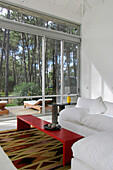 Modernes, helles, weißes Wohnzimmer mit Teppich und rotem Couchtisch und großen Fenstern mit Blick auf den bewaldeten Garten und die Terrasse