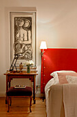 Lit lamp on red headboard in bedroom with modern art above wooden bedside table