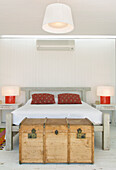 Weiße Pikee-Bettdecke mit roten Kilim-Kissen in einem Schlafzimmer mit Reisekoffer und Hängelampe aus Ripsbändern