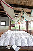 Double bed in glass walled bedroom with swathes of fabric hanging from beams of thatched roof