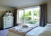 Dolls house on painted chest of drawers in bedroom with large windows and single quilt