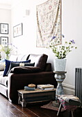 Blumenarrangement auf Vintage-Sockel mit braunem Ledersofa und Wandbehang im Wohnzimmer eines Hauses in London, UK