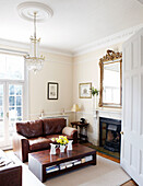 Cut flowers on wooden coffee table with brown leather sofa in living room with gilt-framed mirror in Warwickshire home, England, UK