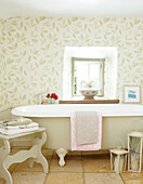 Freestanding bath below window with leaf patterned wallpaper in Devonshire cottage UK