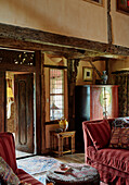 Rotes Sofa mit Wandteppichfußhocker und Blick durch eine Holztür in einem Bauernhaus in Herefordshire, UK