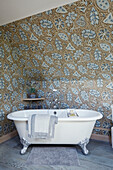 Freestanding clawfoot bath with blue and gold patterned wallpaper in Cotswolds cottage, UK