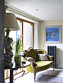 Geknöpftes Sofa am Fenster mit antiker Lampe auf Beistelltisch in einem Bauernhaus in Gloucestershire, England, UK