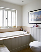 Bathtub below shuttered window in bathroom with nautical artwork in Hampshire farmhouse, England, UK