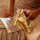 Frau in Abendgarderobe, auf einem Bett sitzend, öffnet eine goldene Stoffhandtasche