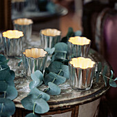 Group of six silver glass tea light candle holders with glowing candles on a rustic side table