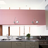 Modern kitchen with aluminium cantilevered wall lights on a striking pink wall over a stainless steel worktop on grey fitted cabinets 