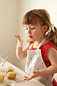 Drei Jahre altes Mädchen steht am Abkühlgitter und bestreut Fairy cakes mit Zuckerstreusel