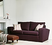 Dunkelviolettes Sofa mit Kissen und Lederfußhocker in einem weiß getäfelten Raum