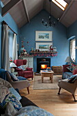 Armchairs and sofa with lit wood burning stove in blue living room of Tiverton farmhouse  Devon  UK