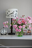 Alarm clock and pink roses with lamp and books on bedside table in Kent home  England  UK