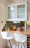 Glass fronted fitted units above wooden kitchen worktop with bar stools in New Malden home, Surrey, England, UK