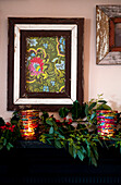 Lit candles on mantlepiece with holly and artwork