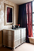 Gold framed mirror above metallic chest of drawers in bedroom of London apartment England UK