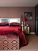 Red velvet bed cover on gold bed with patterned blanket box in bedroom of London apartment England UK