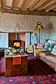 Sofa und Holztruhe mit gemustertem Teppich und beleuchtetem Holzofen im Wohnzimmer eines Hauses in Tregaron, Wales UK