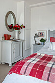 Circular mirror with cut flowers on sideboard in bedroom with red and white checked blanket in Penzance farmhouse Cornwall UK