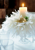 White feather and gold bauble centrepiece with burning candle on a dining table 