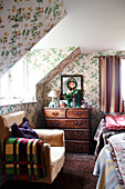 Armchair and wooden chest of drawers below dormer window in floral papered bedroom of Shropshire cottage England UK