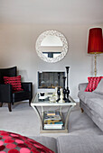 Circular mirror above fireplace with glass topped coffee table and black leather armchair in living room of modern home Bath Somerset, England, UK