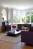 Desk at bay window of living room with purple sofa and low coffee table in London home, England, UK