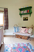 Two seater metal daybed with green wall-mounted bookcase in bedroom of Kent farmhouse England UK