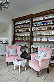 Glassware and crockery on wooden shelves with pair of pink armchairs in Sussex kitchen UK