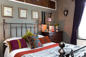 Patterned cushion on wrought iron bed with framed artwork and cupboard storage in Surrey bedroom   England   UK