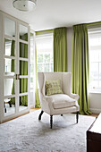 Weißer Sessel und verspiegelter Kleiderschrank mit lindgrünen Vorhängen im Schlafzimmer eines Londoner Stadthauses England UK