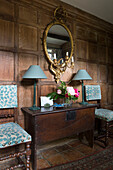 Circular gilt mirror above table with pair of lamps and upholstered chairs in panelled Kent farmhouse UK