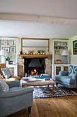 Light blue armchairs with gilt framed mirror above lit fireplace in Gloucestershire cottage UK