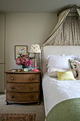 Pink lamp and flowers on wooden chest of drawers at bedside in London home UK