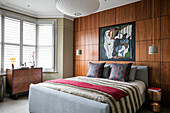 Artwork above double bed with cushions and wood panelling with chest in bay window of London home UK